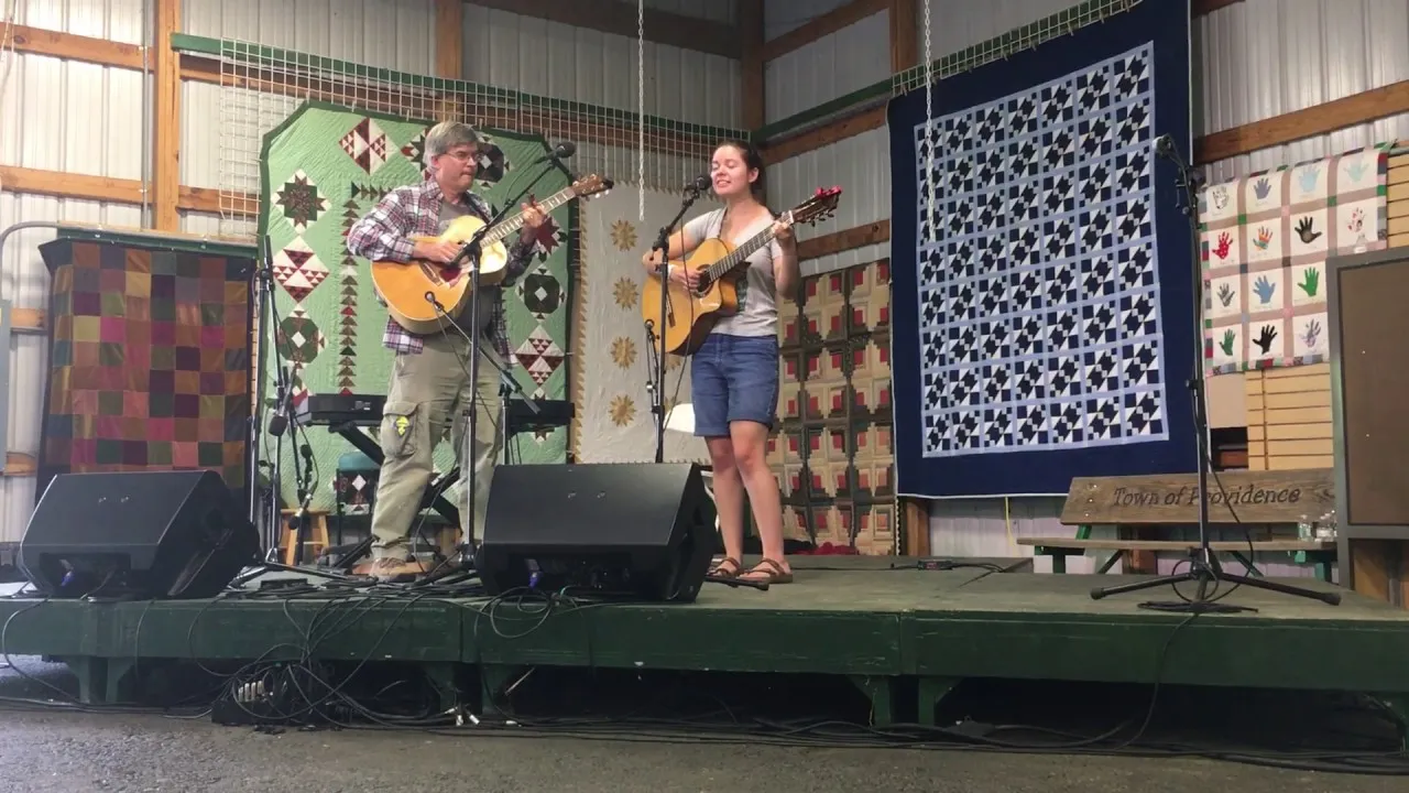 Joe Hetko playing acoustic guitar on stage beside his daughter, who is singing and playing guitar, with quilts hanging behind them.